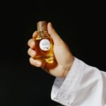 Mão segurando um frasco de perfume árabe dourado contra fundo escuro